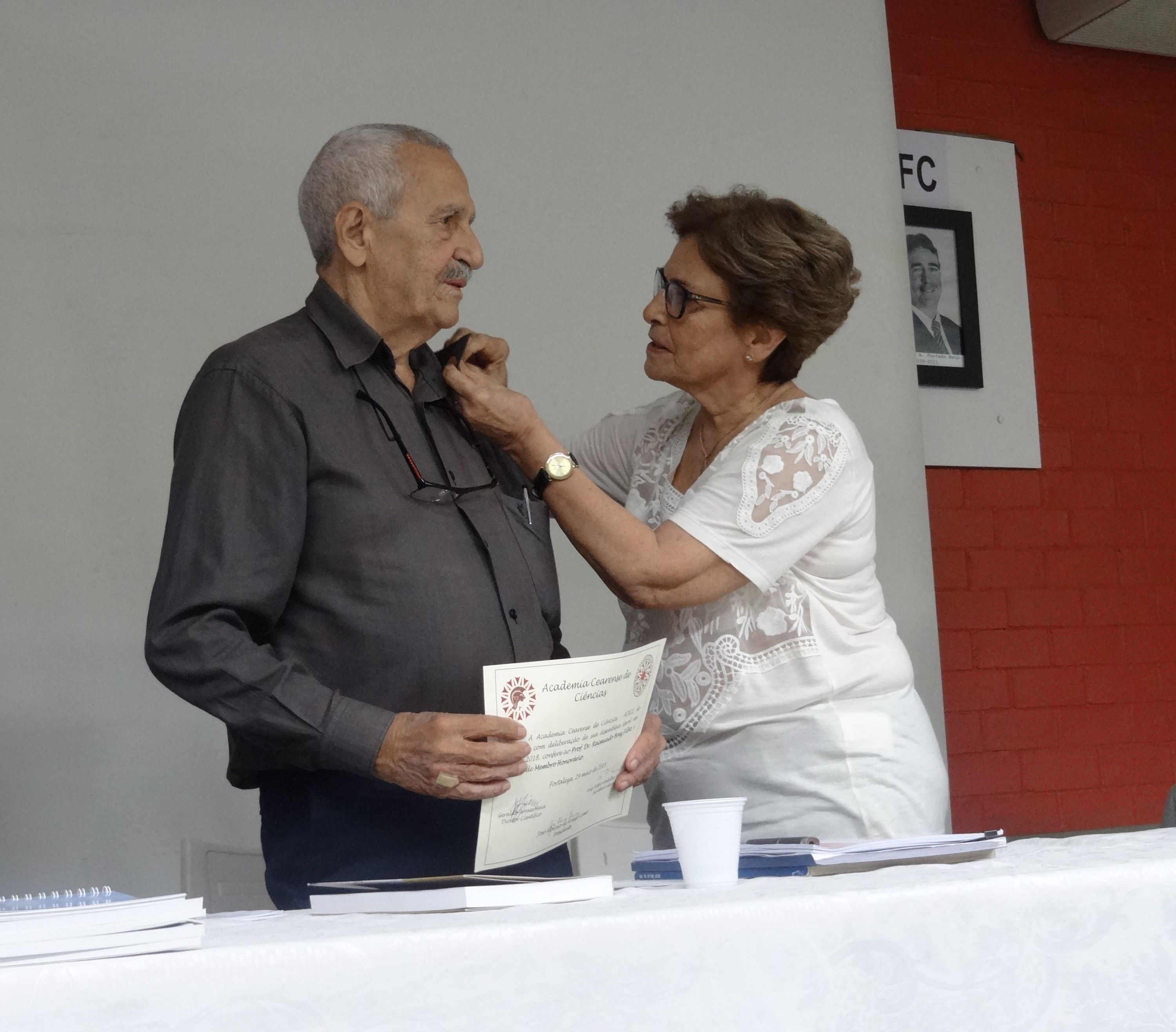 Profa. Glauce Barros Viana colocando o “Botam” da academia na lapela da camisa do agraciado.